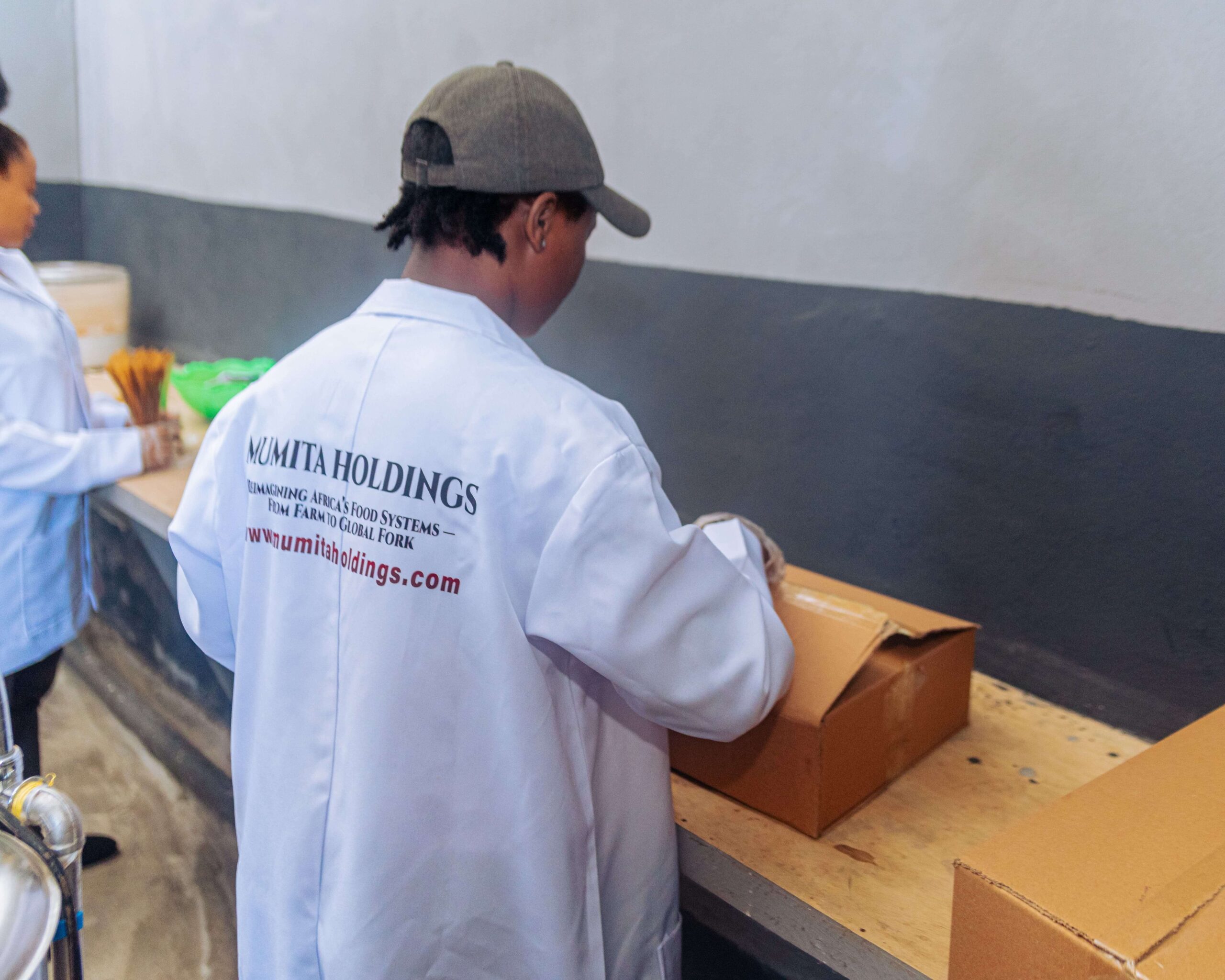 A man in a Mumita Holdings lab coat packs cartons at a bench
