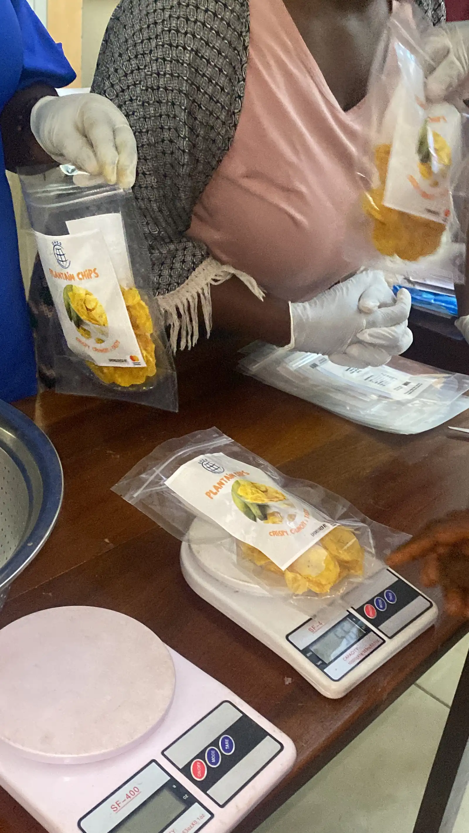 Gloved hands fill a pouch of plantain chips on a scale