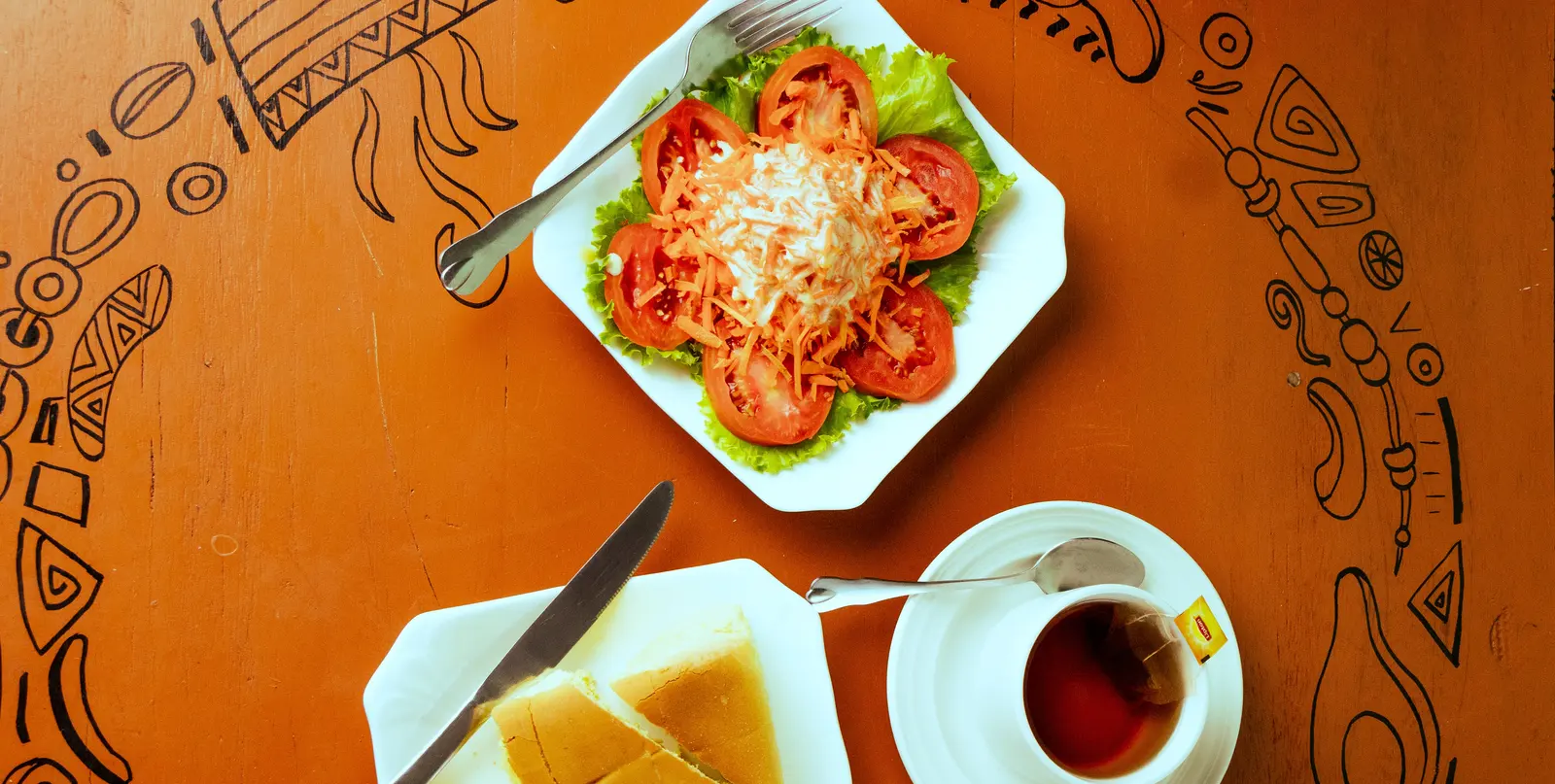 A Praïs salad of tomato, lettuce and grated carrot, with bread and a cup of tea, on a patterned table