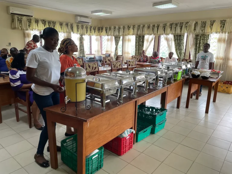 Praïs catering staff behind a long buffet of chafing dishes in a hall, a juice dispenser at the end
