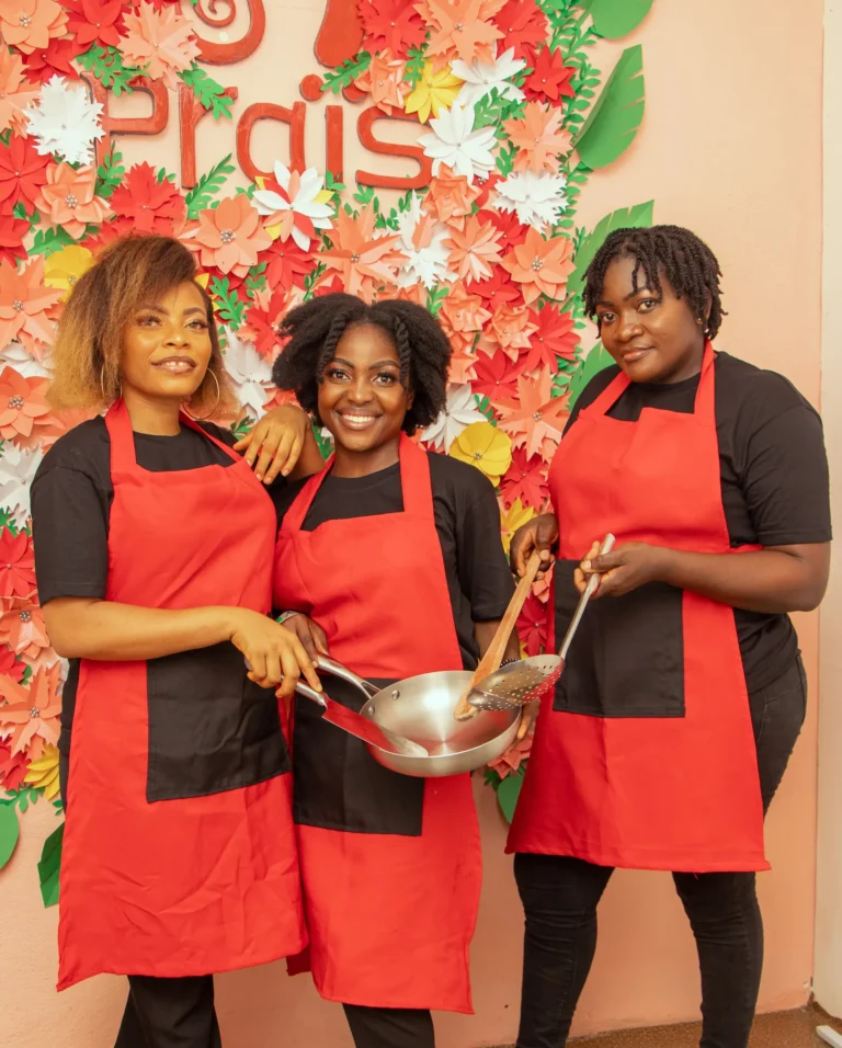 Three Praïs cooks in red aprons mix a bowl of batter in front of the Praïs flower wall