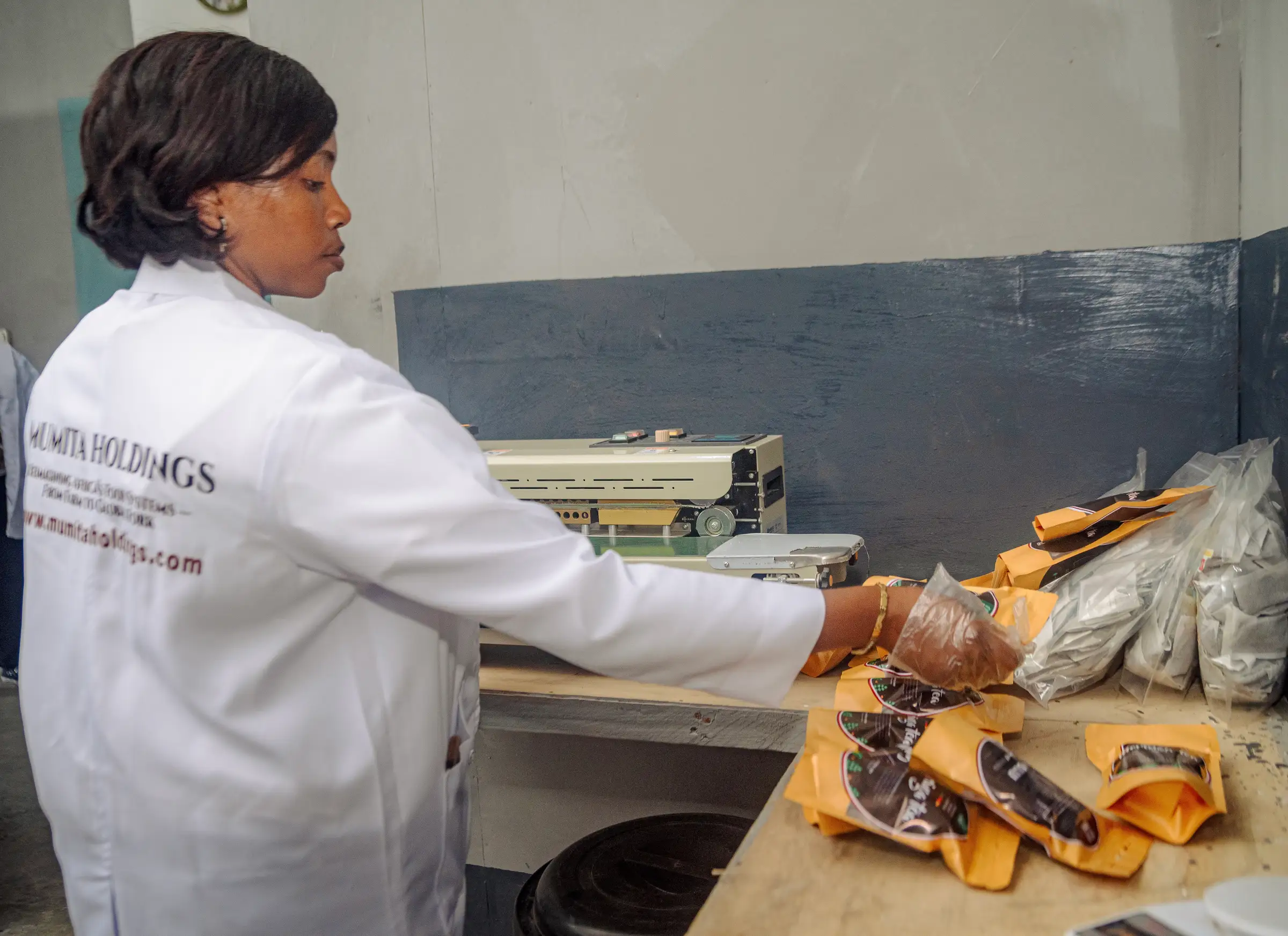 A Mumita staff member in a lab coat seals product pouches beside a band sealer
